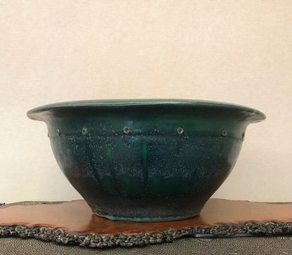 A wide, deep green glazed ceramic pot with speckled surface and small holes around rim.