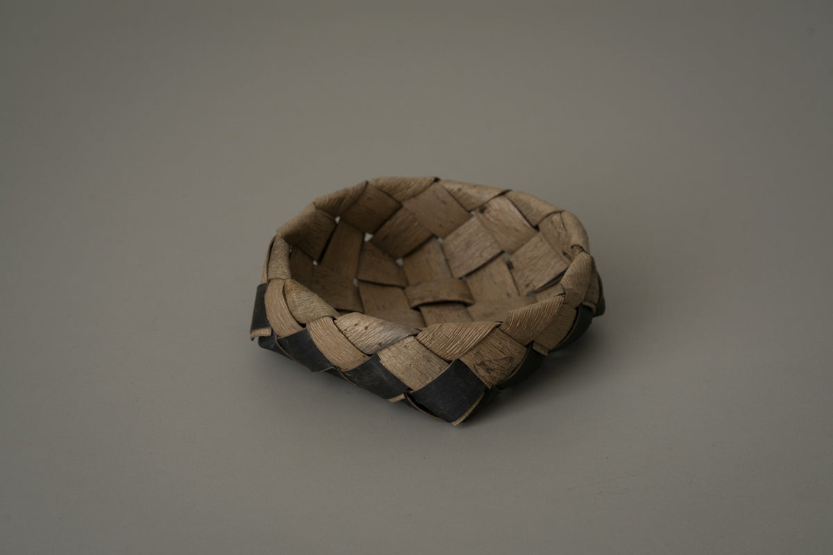 Small shallow box woven from strips of walnut bark in warm brown, on a plain grey ground.