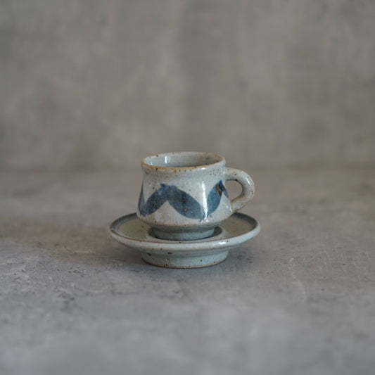 Small white cup painted with blue leaf shapes, standing on a matching blue-rimmed saucer.