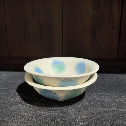 Two cream ceramic bowls stacked one inside the other, painted with soft blue and green pom-pom dots.