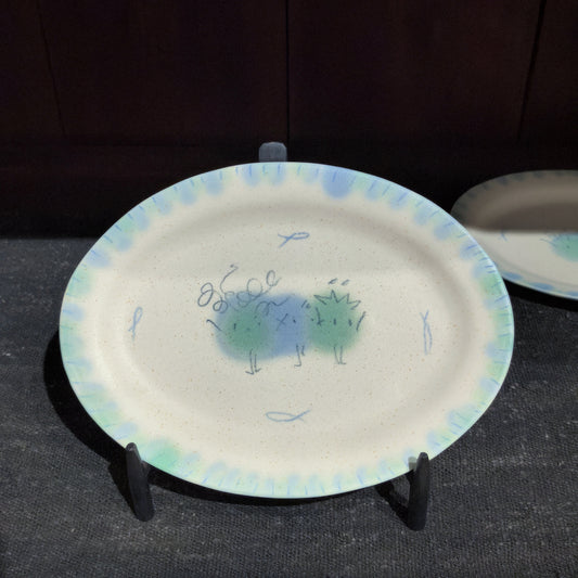 Oval cream ceramic plate with a blue dotted rim and a small green creature painted at its centre.