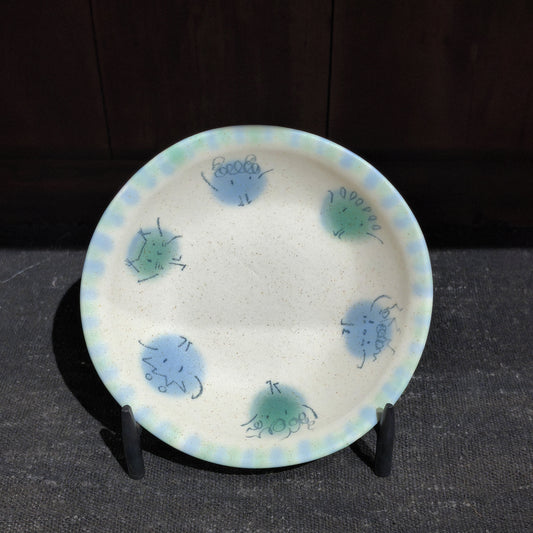 Cream ceramic plate painted with small blue and green pom-pom creatures and a dotted rim, on a black stand.
