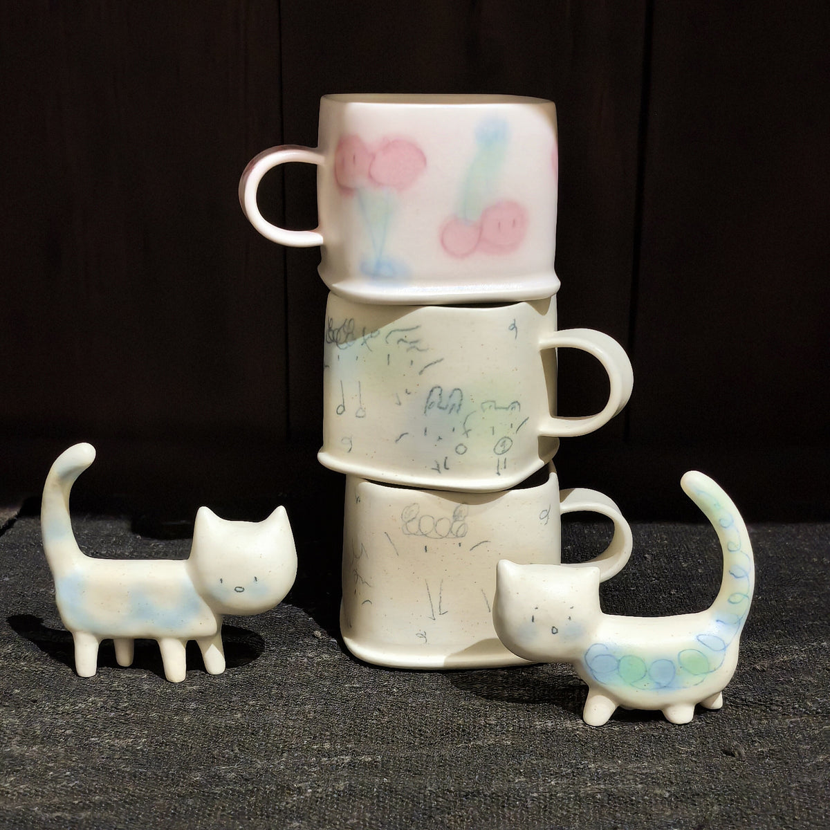 Three cream square cups stacked in a tower with pastel pom-pom marks, flanked by two small ceramic cat figures.