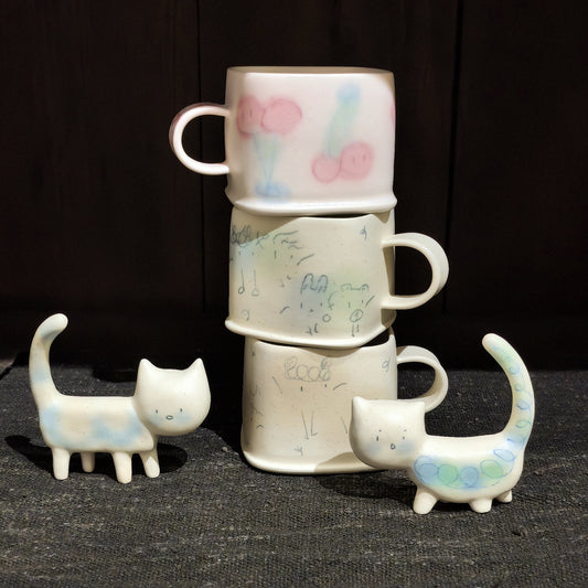 Three cream square cups stacked in a tower with pastel pom-pom marks, flanked by two small ceramic cat figures.