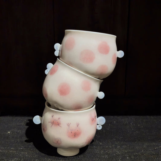 Three cream twin-handled cups stacked in a leaning tower, painted with pink dots and small cat faces.