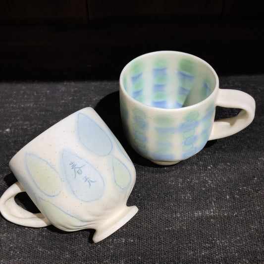 Two cream ceramic cups with open loop handles, painted with a pale blue check pattern and soft yellow ovals.