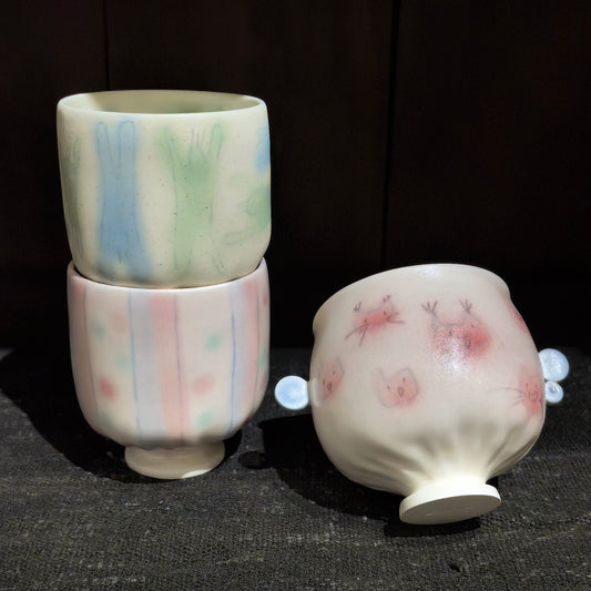 Two cream cups stacked with pastel green and pink brushed stripes, beside a round pink-spotted cup lying on its side.