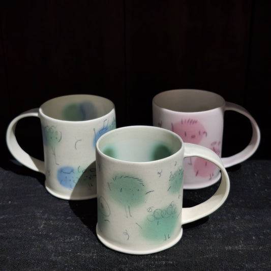 Three cream ceramic mugs with loop handles, each painted with a fluffy green or blue pom-pom creature.
