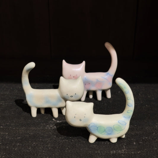 Three small ceramic cat figures with long curved tails, cream with soft pink and blue markings.