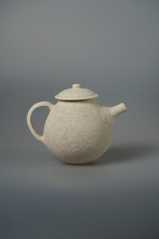 Round cream teapot with a coarse sandy surface, a knopped lid and a curved handle.