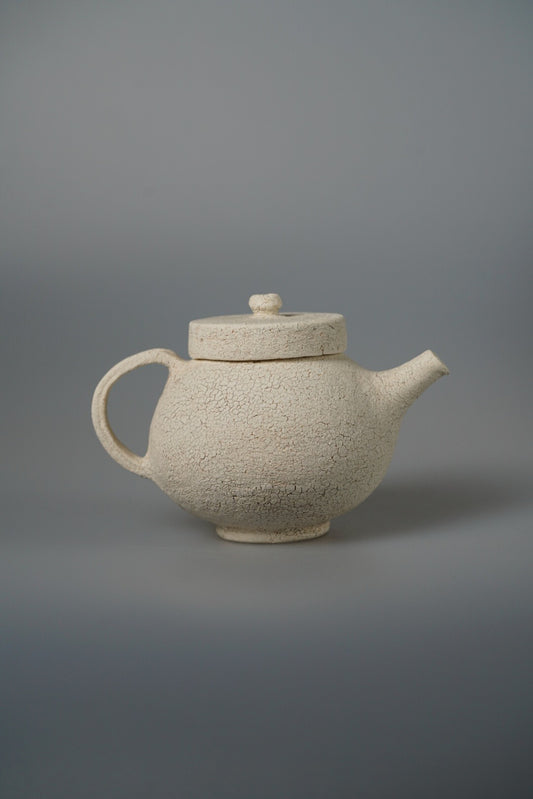 Cream teapot with a flat disc lid and a broad rounded body, on a pale grey ground.