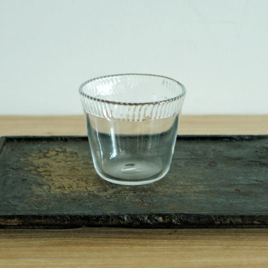Short clear glass cup with a ribbed upper band and a smooth base, on a dark weathered board.