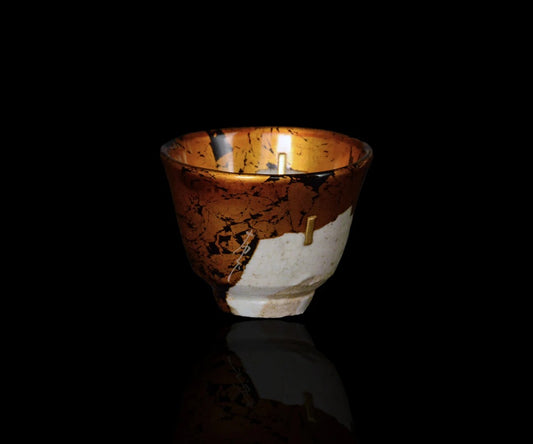 Small cup lit against black, its dark surface broken by broad panels of gold and pale lacquer repair.