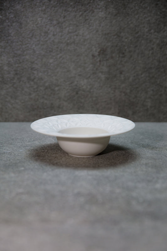 Shallow white porcelain bowl with a wide flat rim cut in a fine radiating pattern, on a grey ground.