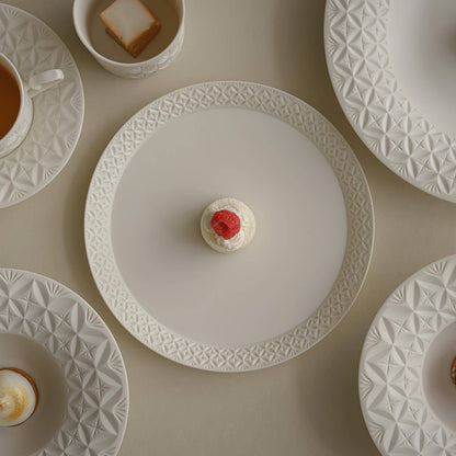 White porcelain plates cut with fine geometric bands, seen from above beside cups and a small pastry.