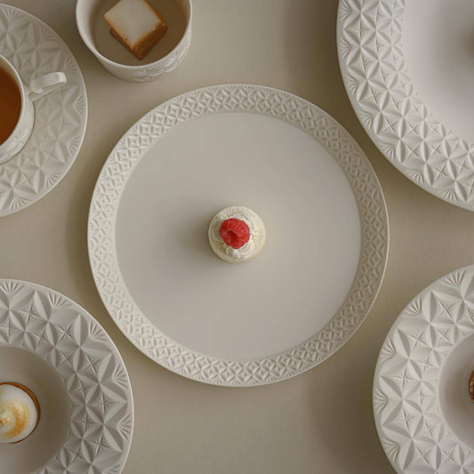 White porcelain plates cut with fine geometric bands, seen from above beside cups and a small pastry.