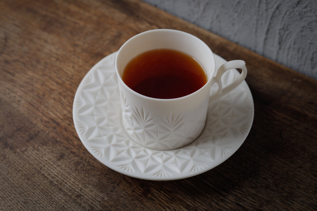 White porcelain cup of dark tea on a matching saucer, both cut with a fine faceted pattern, on a wooden table.