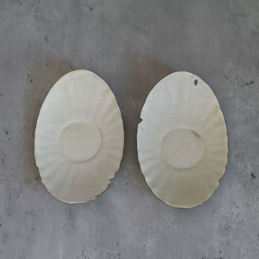 Two oval white enamel coasters with fluted edges and a shallow round well, on concrete.