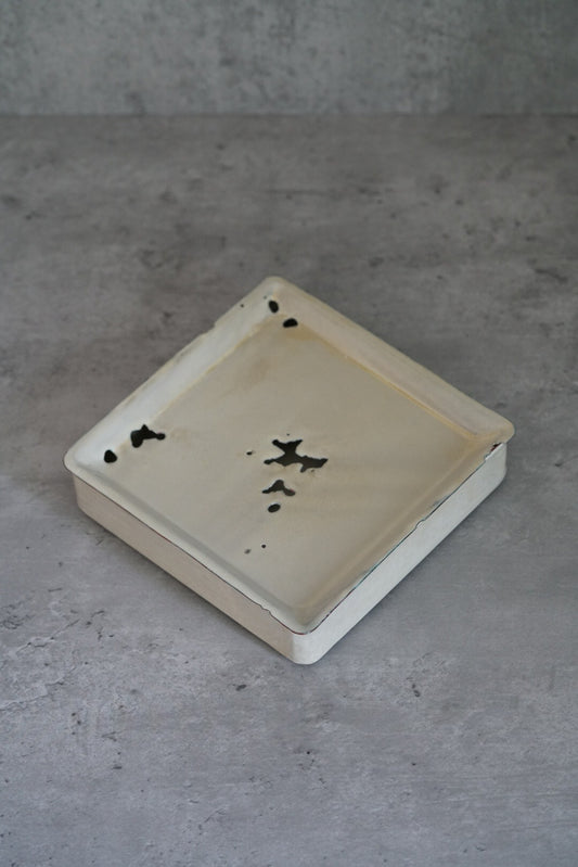 Square white enamel tea stand with a raised rim and small dark flecks across its face.