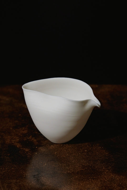 White porcelain pouring bowl with thin walls folded to a sharp pinched spout, on a dark surface.