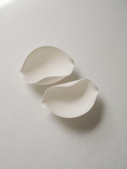 Two curved white porcelain scoops shaped like open petals, lying on a pale textured cloth.