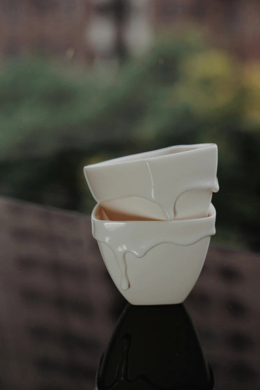 Two white porcelain cups stacked outdoors, their rims folded and drooping like the top of a milk bottle.