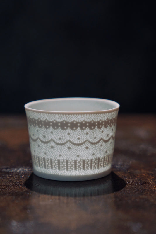 Straight-sided white porcelain cup banded with a delicate pierced lace pattern, on dark wood.