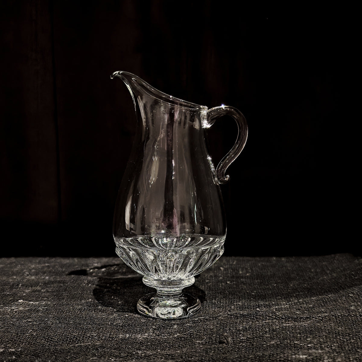 Clear glass pourer with a drawn lip and curled handle, standing on a faceted foot on dark wood.