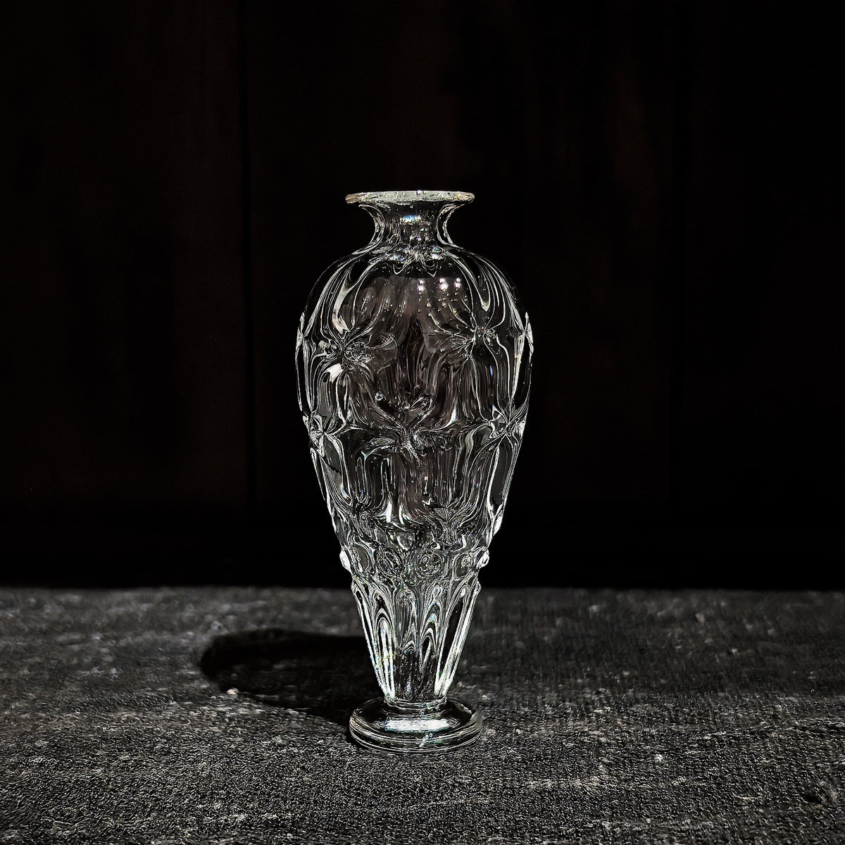 Slim clear glass vase with a narrow neck and a body cut in soft vertical ribs, on dark wood.