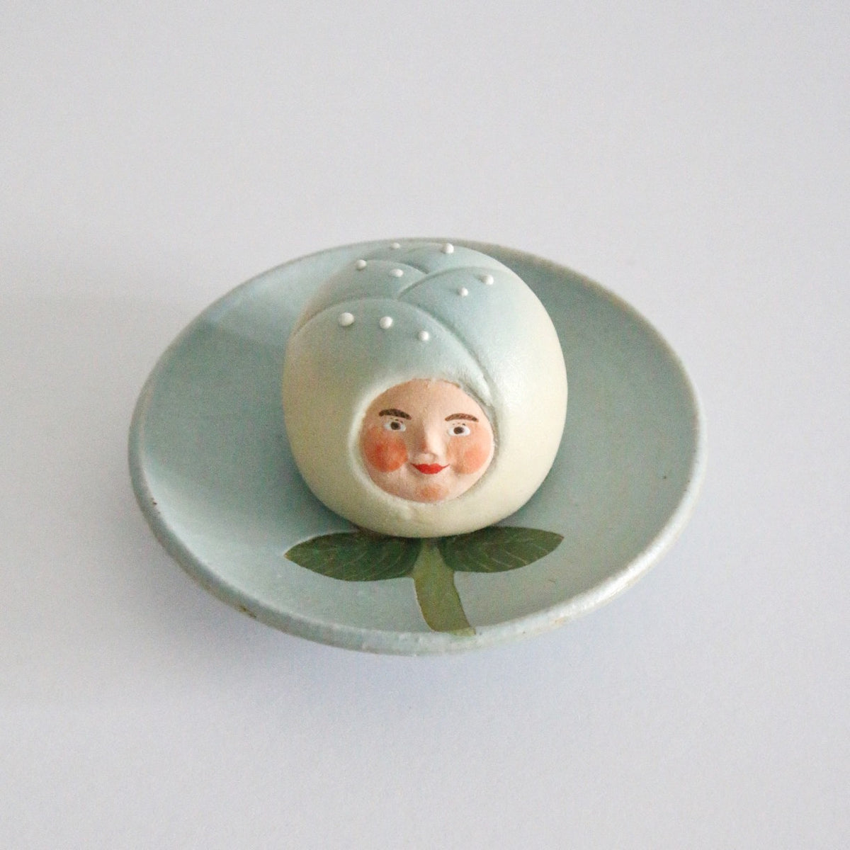Ceramic sweet with a pale blue dotted hood and a painted face, on a pale green leaf dish.
